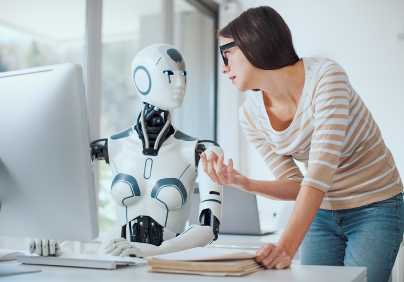 A woman speaks to a robot at a computer, illustrating a modern dialogue between llinkedin Writer and artificial intelligence.